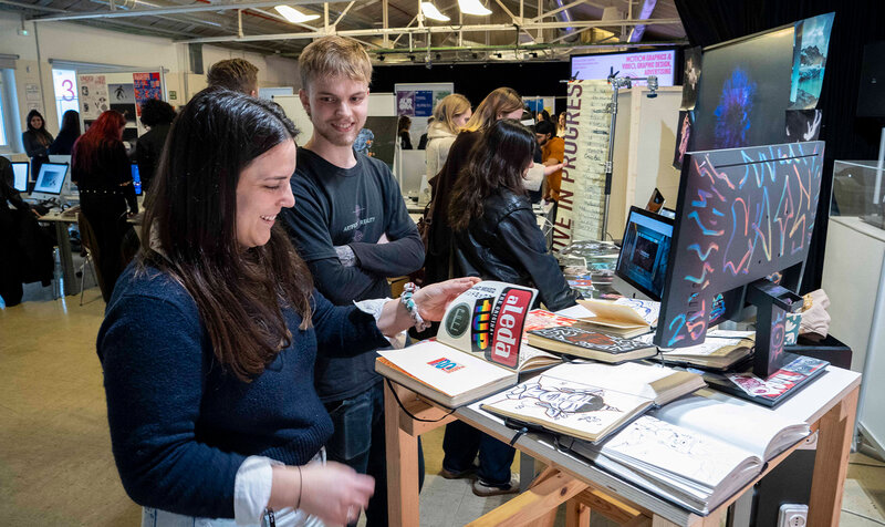 A woman browses magazines in the foreground at a graphic design exhibition with more people in the background