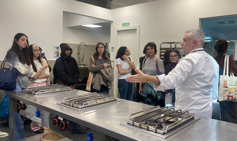 University students in a professional kitchen listening to a chef's explanations