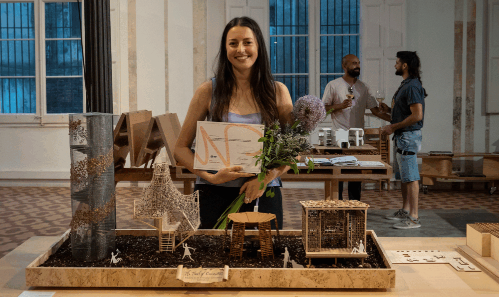A girl with a bouquet of flowers and a diploma in her arms at an exhibition in front of an interior design model. In the background, two people are chatting.